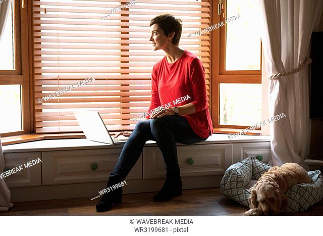 Woman looking through window while using laptop in living room