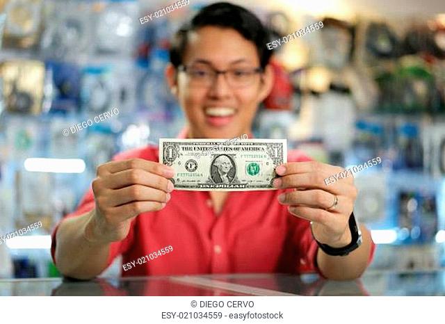 Young asian shop owner in computer store, showing his first dollar to the camera and smiling. Focus on banknote