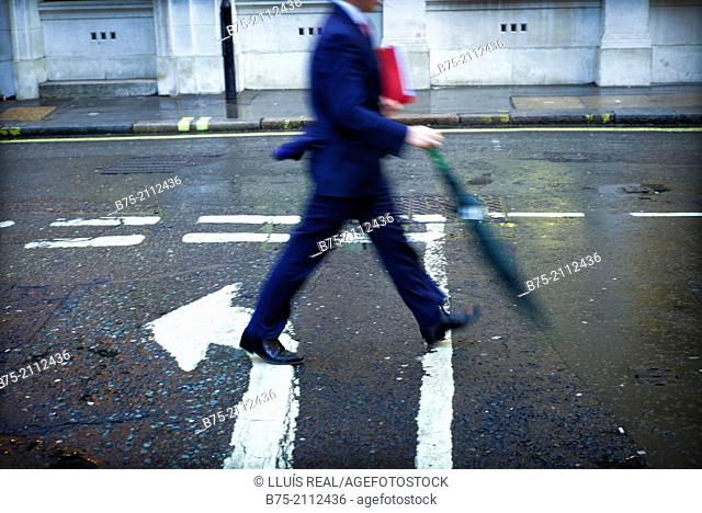 unrecognizable executive man, with a folder under his arm and an umbrella walking down the street in the opposite direction of an arrow painted on the asphalt