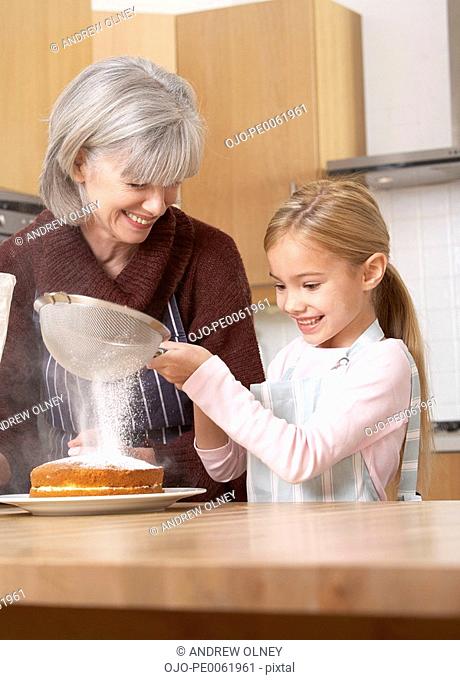 Woman and young girl in kitchen sifting icing sugar onto a cake and smiling