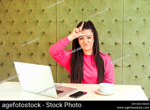 Negative rude young girl with black dreadlocks hairstyle in pink blouse is sitting in cafe and having bad mood, showing loser sing with finger and looking to...