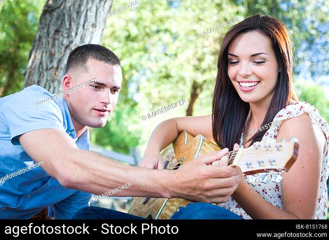 Young adult man teach his girlfriend how to play the guitar outside in the park