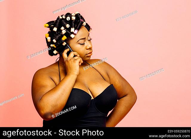 Woman with hair curlers and neglige talking on the phone with eyes closed, isolated on pink background