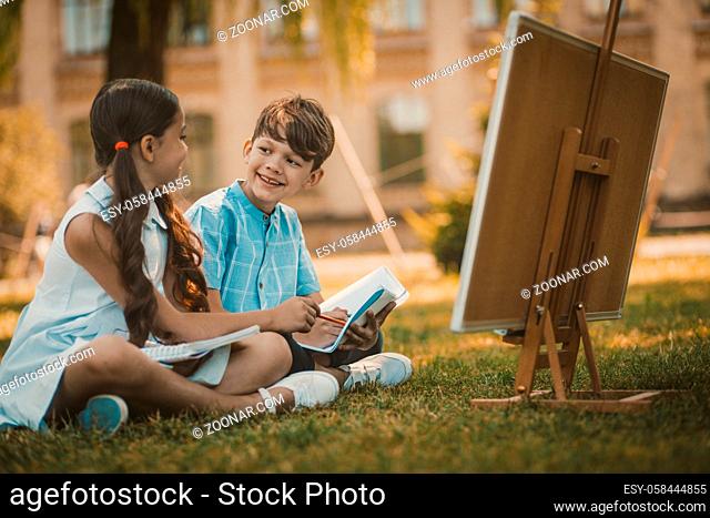 Best Friends Kids Sitting At Grass In Park, Cheerful Children, Boy And Girl In White Dress Looks Each Other Holding Copybook, Artboard Are Nearby, Toned Image