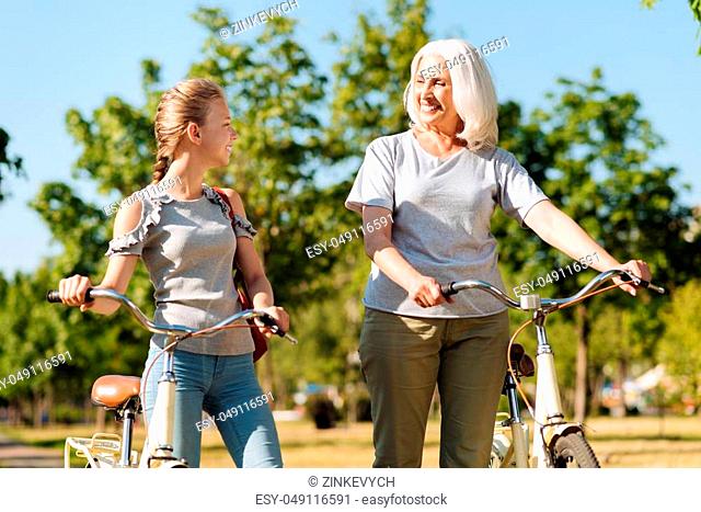 Pleasant time together. Joyful little girl and her positive grandmother talking and standing in the park while resting after riding a bicycle