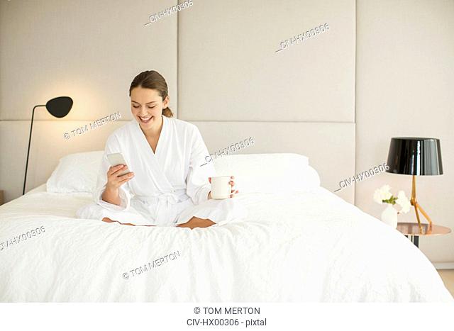 Smiling woman in bathrobe drinking coffee and texting on cell phone on bed