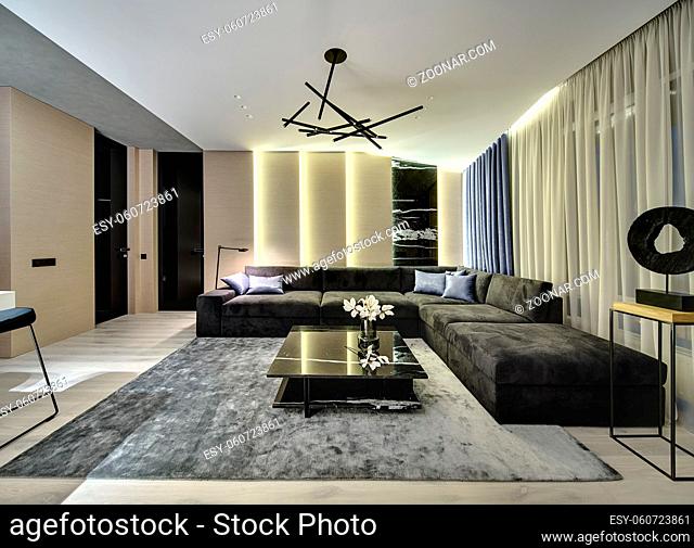 Room in a modern style with wooden walls with a marble part. There is a dark sofa, black marble table with flowers, gray carpet, large window with curtains