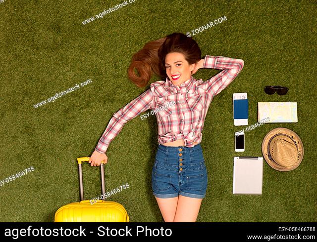 Happy beautiful tourist lady lying on green grass and holding yellow luggage. Pretty woman with red lips lying among mobile or smart phone, passport, cap