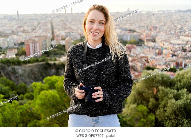 Portrait of smiling young woman with camera at sunrise above the city, Barcelona, Spain