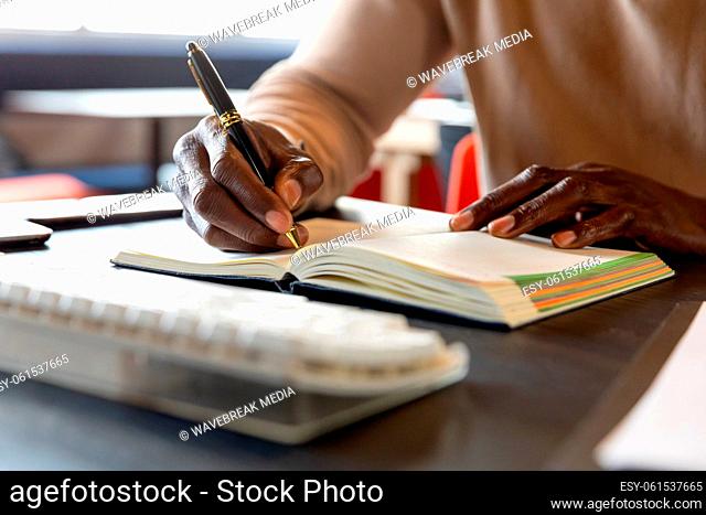 Midsection of african american mature businessman writing in diary at desk in office