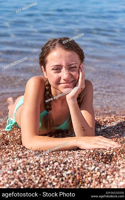 A teenage girl with freckles on her face and pigtails lies on the beach near the sea. Summer sunny day. Portrait and look into the camera