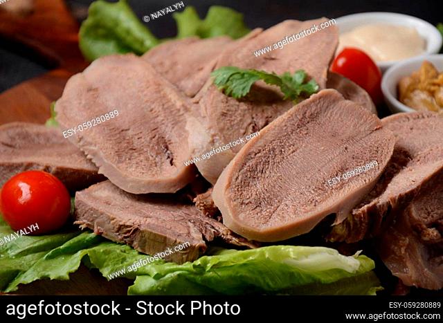 Sliced Beef Tongue Slices on a platter with lettuce leaves, cherry tomatoes and Dijon mustard on a wooden background
