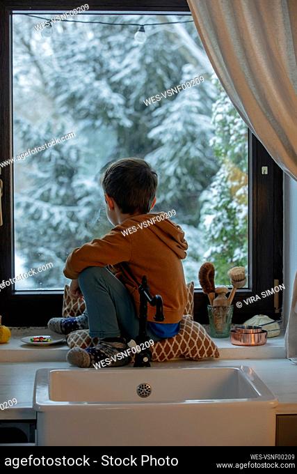 Boy sitting on window sill looking out in kitchen at home