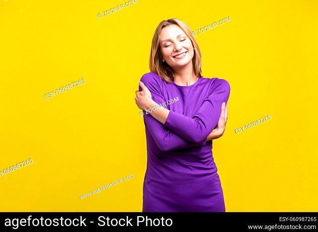 I love myself. Portrait of satisfied selfish woman in elegant tight purple dress embracing herself, standing with closed eyes smiling from pleasure