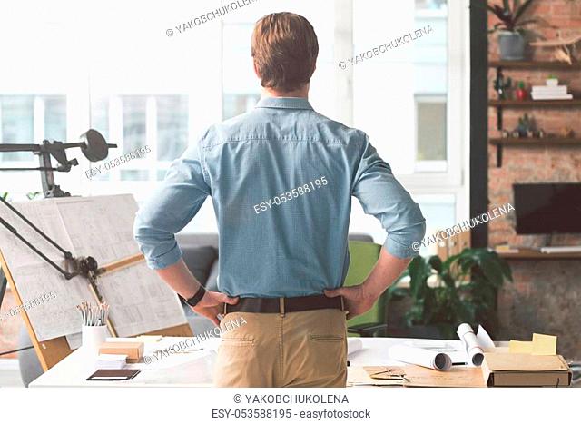 Back view of elegant young man is resting in office with arms akimbo. He is standing in front of workplace and looking through window