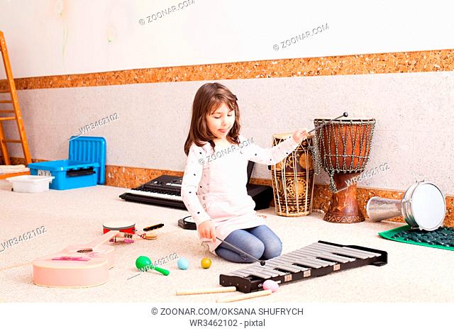 The cute little girl sits on the floor and playing on the xylophone at the kindergarten. The girl is testing various musical instruments