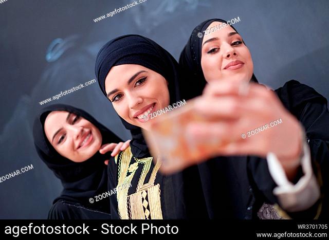 group of young beautiful muslim women in fashionable dress with hijab using mobile phone while taking selfie picture in front of black chalkboard representing...