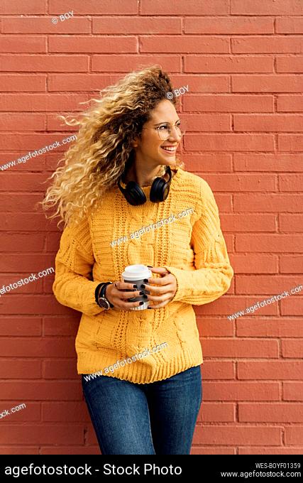 Beautiful young blond woman with long curly hair looking away while holding disposable coffee cup against red brick wall