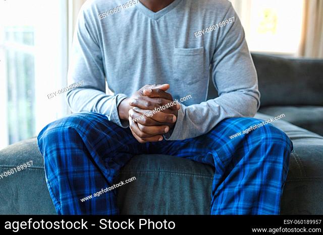 Midsection of african american man with hands clasped, thinking and sitting by window in living room