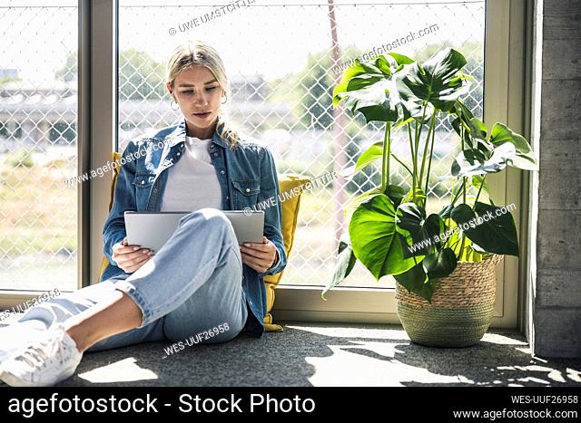 Businesswoman using tablet PC sitting on floor by plant in office