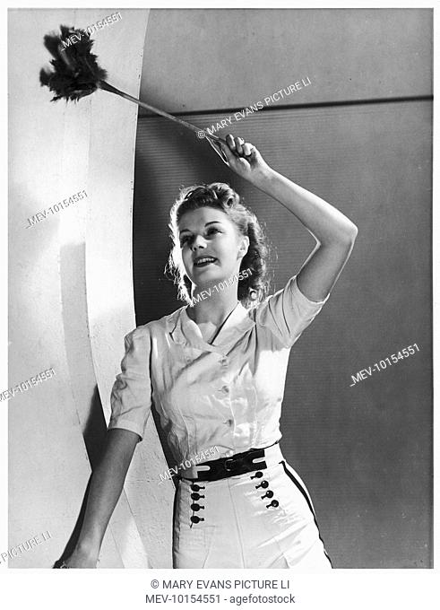 A smiling young lady in shorts with buttoned falls dusts briskly with a feather duster - a great way to tone up the waist and bust without the need of a gym