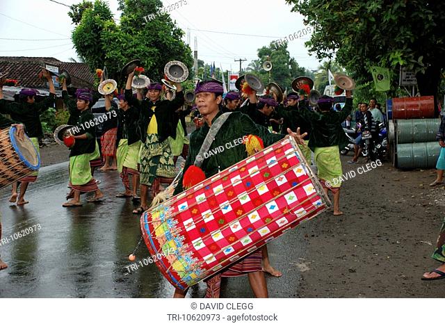 Sasak man in traditional dress of black jacket and sarong playing large colourful drum leads other musicans through wet village street in Muslim wedding...