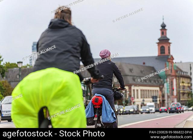 PRODUCTION - 28 August 2023, Hesse, Frankfurt/Main: Two people ride their bikes across the Old Bridge, with a view of the Church of the Teutonic Order