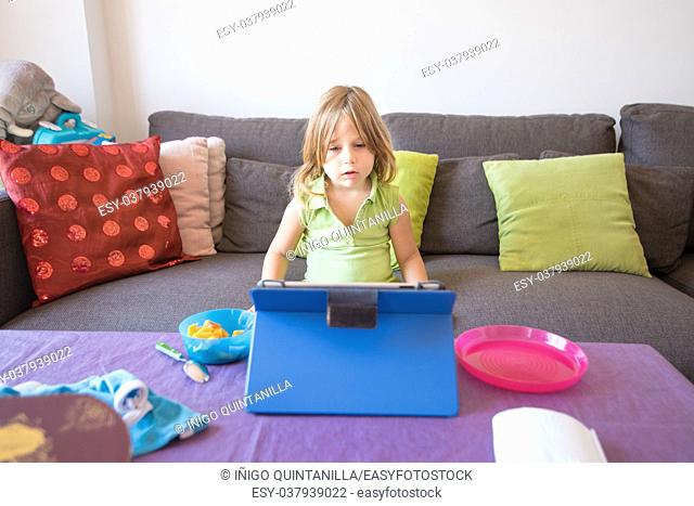 four years old blonde child with green sleeveless shirt sitting on brown sofa, watching digital tablet on table