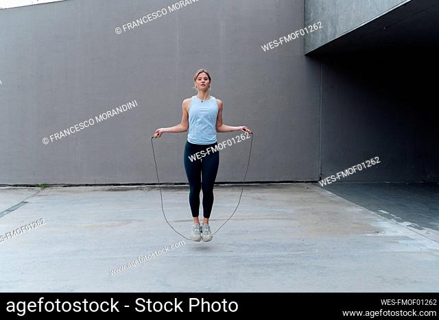 Young sportswoman skipping rope while standing against wall
