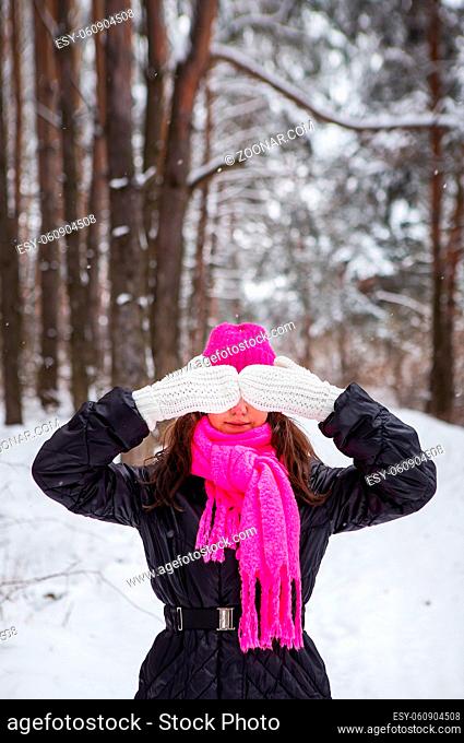 A girl in a pink scarf and hat stands in the middle of a pine forest and covers her face with white mittens. Beautiful pine forest on a frosty day