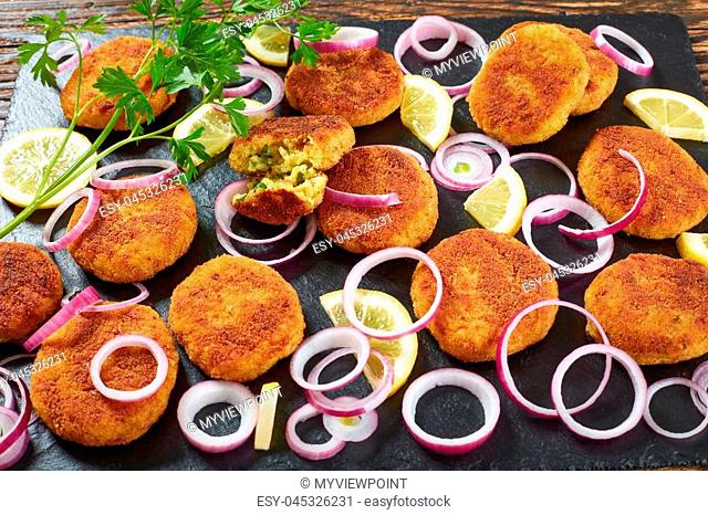 delicious crispy rice patties with crumbled paneer cheese and finely chopped greens on a black slate plate with lemon slices and onion rings on wooden table