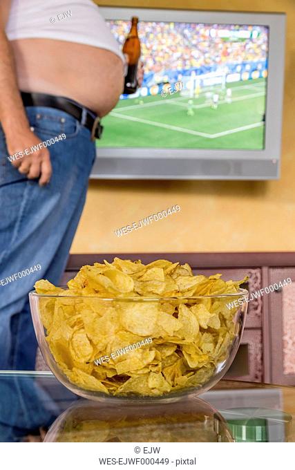 Man with naked beer belly holding beer bottle in front of flatscreen TV broadcasting a soccer match, partial view