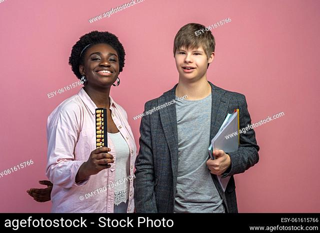 Homeschooling. Dark-skinned woman with abacus and caucasian guy with down syndrome holding notebook smiling at camera standing on light background