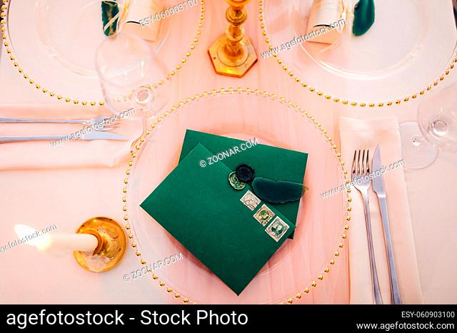 Blank green envelopes with stamps lie on a transparent plate on a set table. A lighted candle is burning on the left. Top view. High quality photo