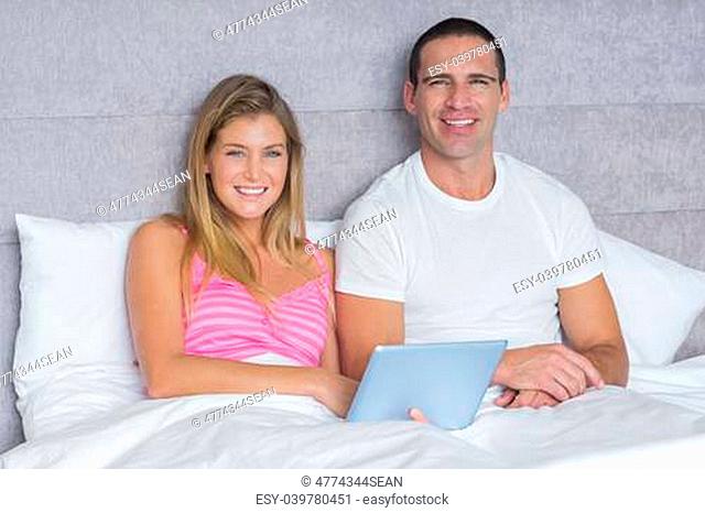 Happy young couple using their tablet pc together in bed looking at camera in bedroom at home