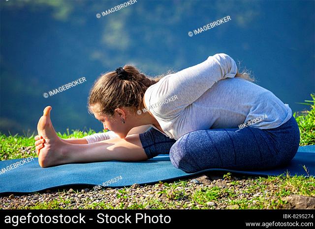Woman doing Ashtanga Vinyasa yoga asana Ardha baddha padma paschimottanasana half bound lotus intense west stretch poser outdoors in mountains in the morning in...