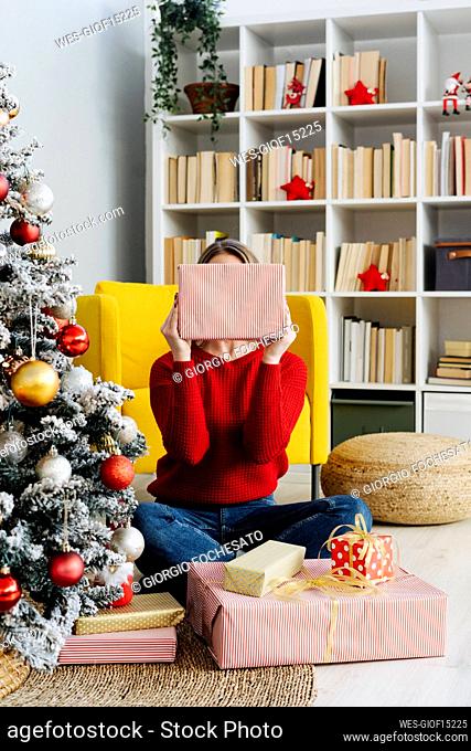 Smiling woman covering face with Christmas present in living room at home