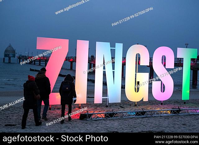28 December 2021, Mecklenburg-Western Pomerania, Zingst: A colorfully illuminated lettering is set up in the evening at dusk in front of the pier on the beach...