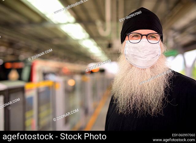 Portrait of mature bearded hipster man as tourist with mask for protection from corona virus outbreak at the sky train station