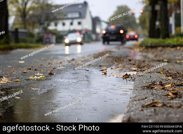 05 November 2023, North Rhine-Westphalia, Duisburg: Raindrops fall into a puddle next to a road while cars drive with their headlights on