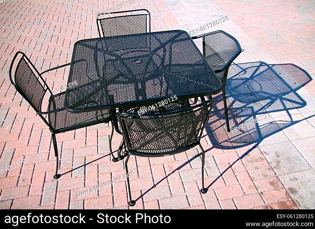 An outside iron table with four iron chairs with their shadow on a concrete floor outdoors