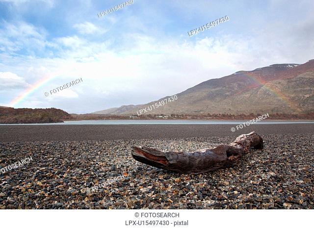 Driftwood log on pebble shore of Lago Grey with full rainbow arching across blue sky, Torres del Paine, Southern Chile, Patagonia
