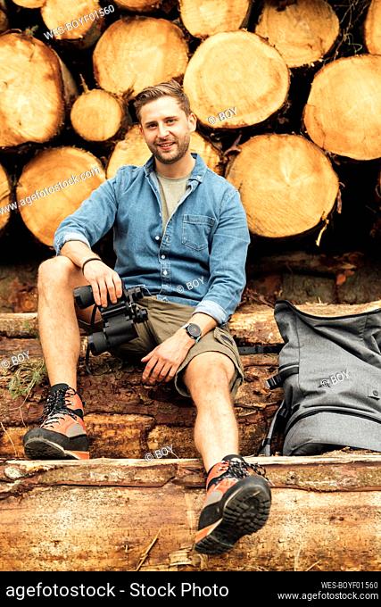 Smiling man with binoculars and backpack sitting on log against woodpile