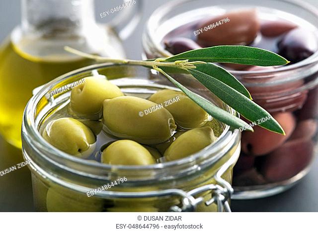 Pickled green and black olives in glass jar with branch leaves close up