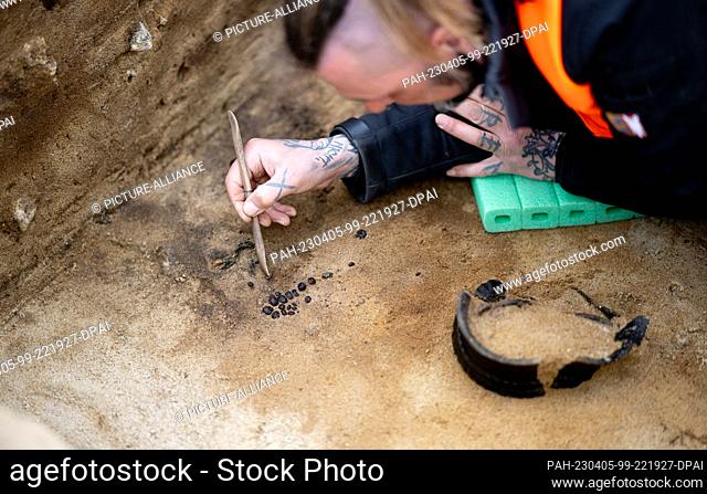 05 April 2023, Schleswig-Holstein, Lohe-Rickelshof: A man uncovers a pearl necklace and a decorated ceramic pot in a grave