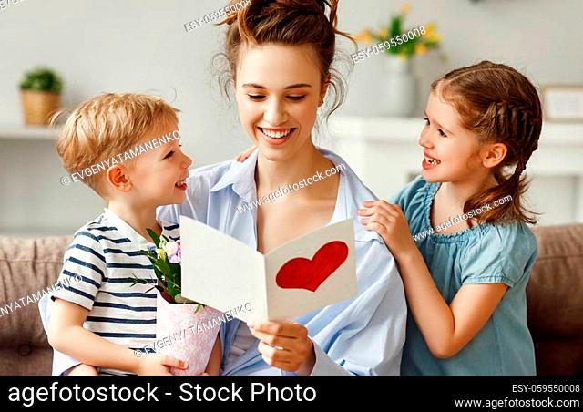 Pleased mother and little children with bouquet of fresh flowers embracing on sofa while reading postcard with red printed heart at home