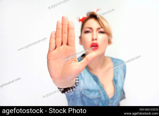 Do not come to me. Portrait of serious beautiful young woman in casual blue denim shirt with red headband standing and looking at camera with stop gesture