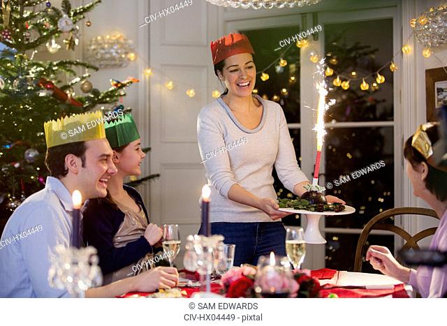 Smiling woman in paper crown serving Christmas pudding with fireworks to family at dinner table