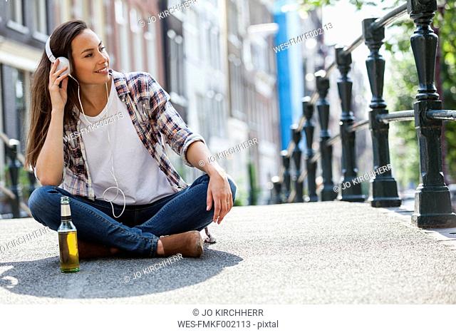Netherlands, Amsterdam, young woman sitting with headphones and beer bottle on bridge