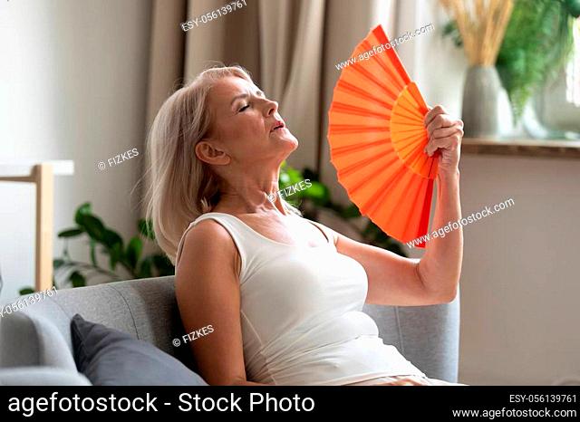 Stressed annoyed old senior woman using waving fan suffer from overheating, summer heat health hormone problem, no air conditioner at home sit on sofa feel...